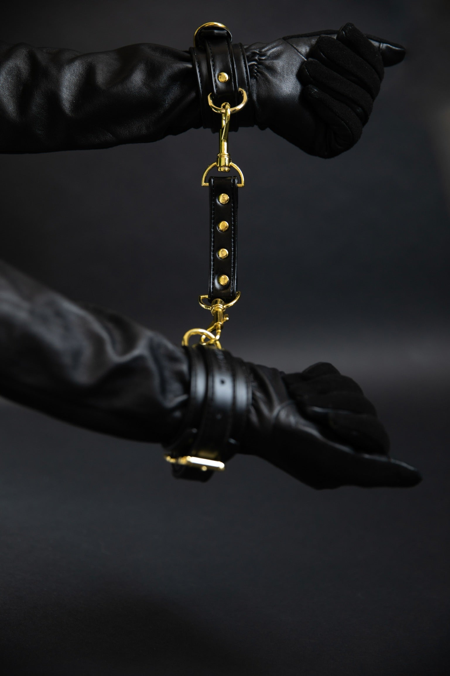 Gloved hands gently joined by black leather cuffs with a connecting band, photographed against a dark background as part of Dahlier’s Eros Collection.