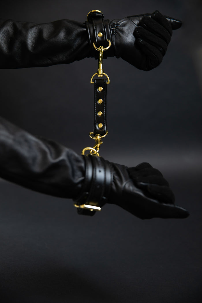 Gloved hands gently joined by black leather cuffs with a connecting band, photographed against a dark background as part of Dahlier’s Eros Collection.